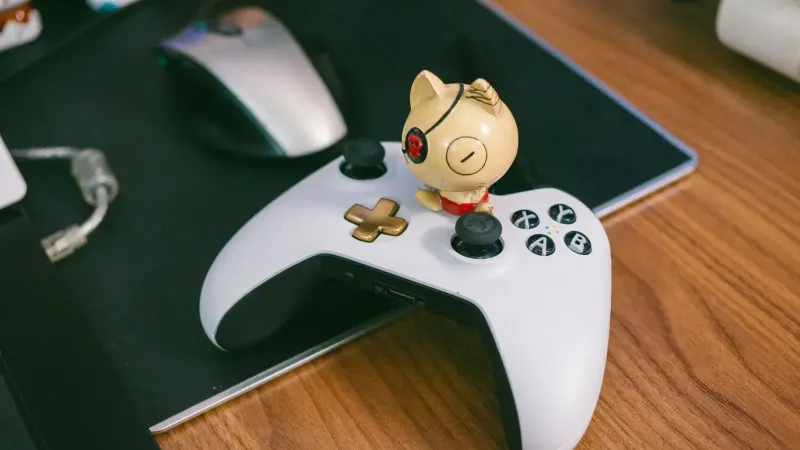 A charming cat figurine sits atop a gaming controller, beside a computer mouse, on a wooden desk.