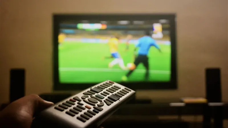 Close-up of a hand holding a remote control in front of a TV showing a football match.