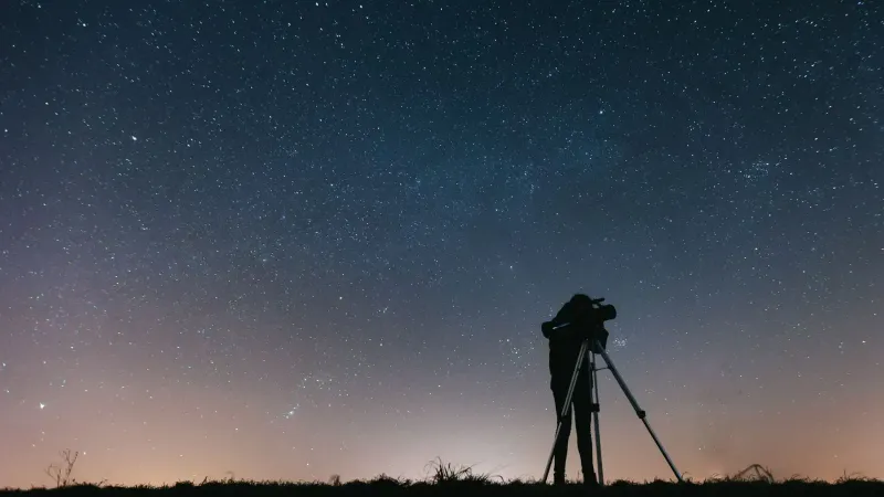 A silhouetted person using a telescope to observe the night sky filled with stars.
