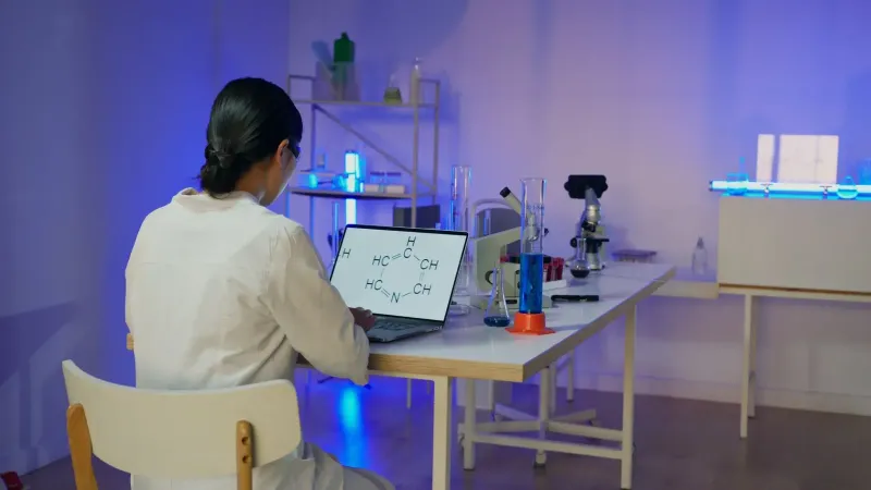 Woman in lab coat analyzing chemical formula on laptop in modern laboratory setting.