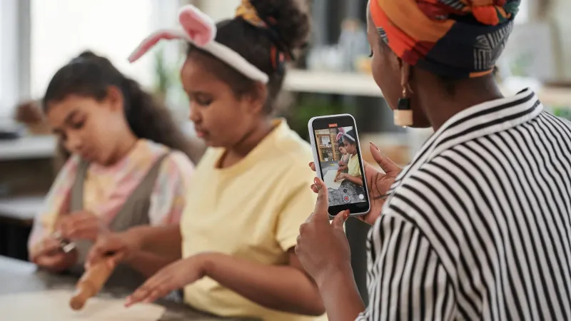 A mother records her daughters baking at home with a smartphone, capturing fun family moments.