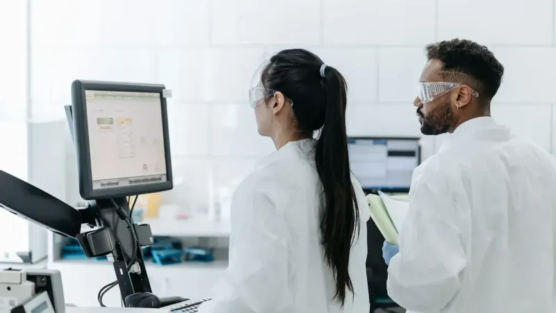 Two scientists wearing lab coats and goggles analyze data on a computer in a modern laboratory.