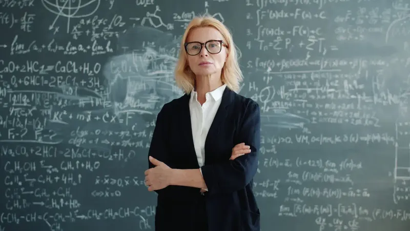Female professor stands confidently with folded arms in front of a chalk-drawn chalkboard filled with equations.
