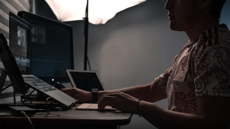 Un hombre editando fotos en múltiples pantallas en un escritorio con poca iluminación.