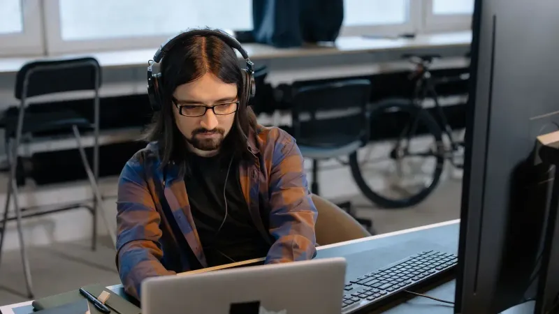 Desarrollador masculino barbudo en auriculares que codifican en una computadora portátil en un entorno de oficina moderno.