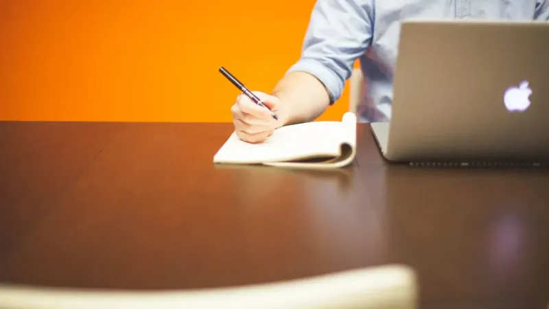Adultos escribiendo notas en un cuaderno junto a una computadora portátil en un escritorio de madera con fondo naranja.