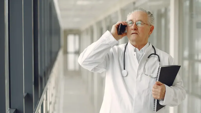 Serious mature male doctor in medical uniform standing with stethoscope and notebook while talking on smartphone in clinic corridor