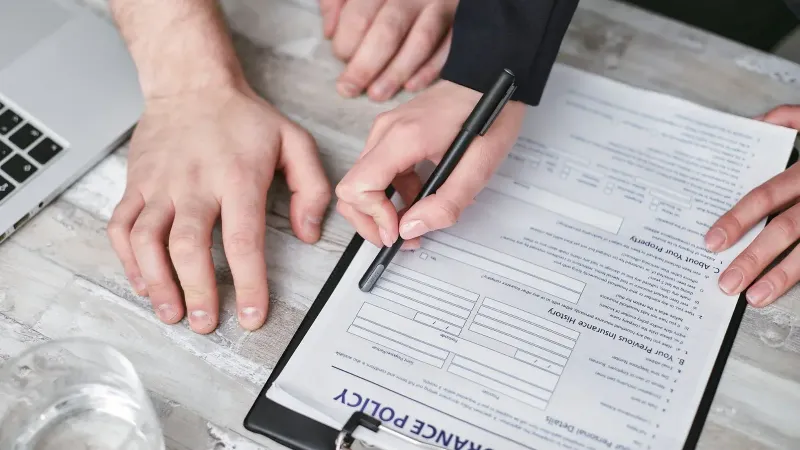 Imagen de cerca de dos personas firmando un documento de póliza de seguro en un escritorio de madera.
