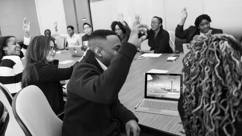 A diverse group of adults in an office meeting, engaged in a discussion with laptops, raising hands to vote onsomething.
