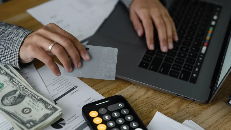 Close-up of a person using a credit card and laptop in a financial setting with cash and calculator.