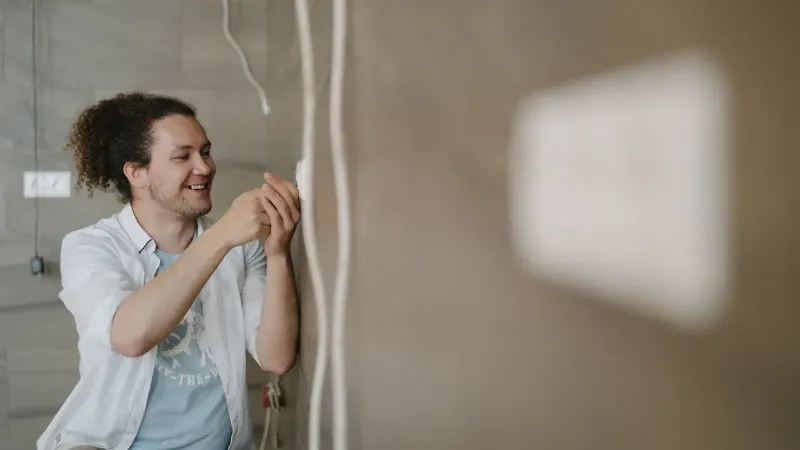An electrician with curly hair adjusts an outlet indoors, smiling as he works.