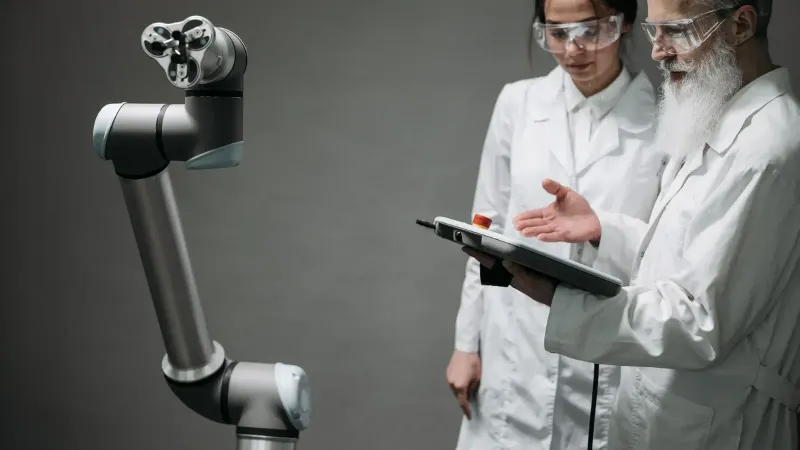 Researchers in lab coats and safety glasses engaging with a robotic arm in a lab setting.