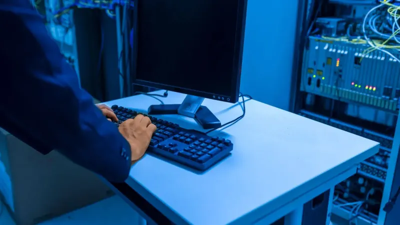 An IT professional operates a computer in a server room, managing network systems and connected devices.