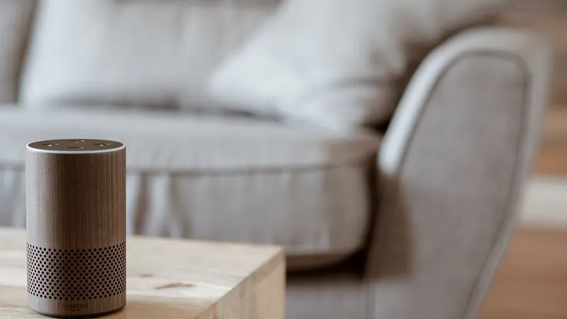 A modern smart speaker sits on a wooden table in a cozy, minimalist living room setting.