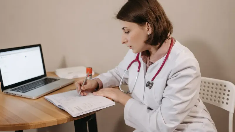 A female doctor in a lab coat writing a prescription in a clinic office setting with a laptop on the table.