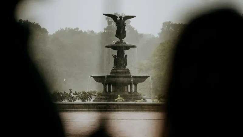 A beautiful angel statue fountain in a lush park during rainfall, creating a serene ambiance.