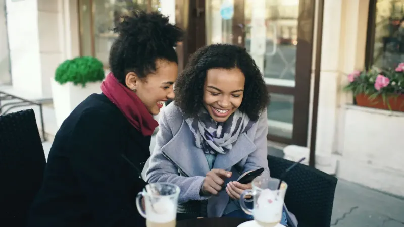Dos amigos riendo tomando un café en un café al aire libre, compartiendo un momento de alegría.