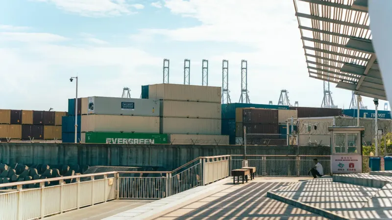 Vista de un puerto de envío con contenedores apilados y un hombre descansando cerca bajo sombras industriales.