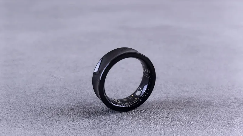A modern black smart ring with visible circuitry resting on a textured concrete surface.