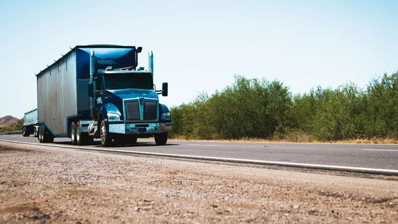 A blue semi truck driving on a rural highway under clear blue skies, showcasing transportation and travel.