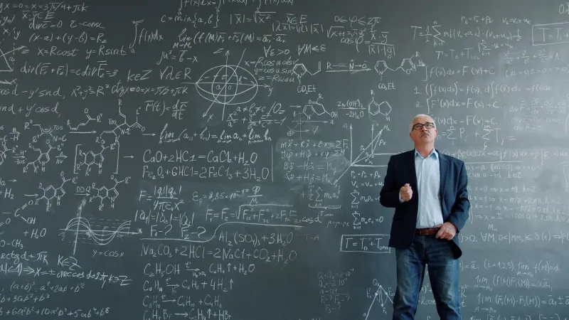 Professor standing in front of a blackboard filled with complex equations and scientific formulas.