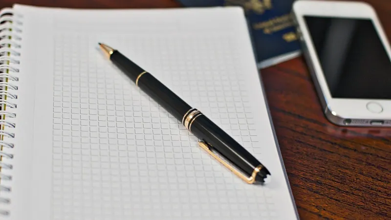 Black pen on a grid notebook with smartphone and passport on wooden desk.