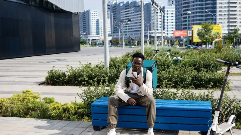 Young delivery man on a break with electric scooter and thermal bag in modern cityscape.