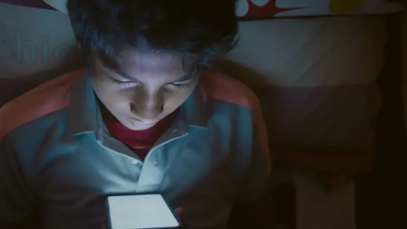 Boy lying in bed staring intently at a glowing smartphone screen.