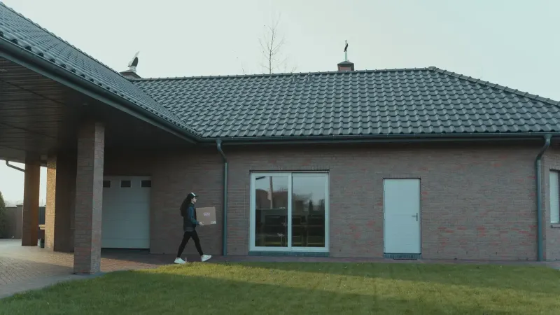 Person delivering package to a modern brick house with a tile roof and green lawn.