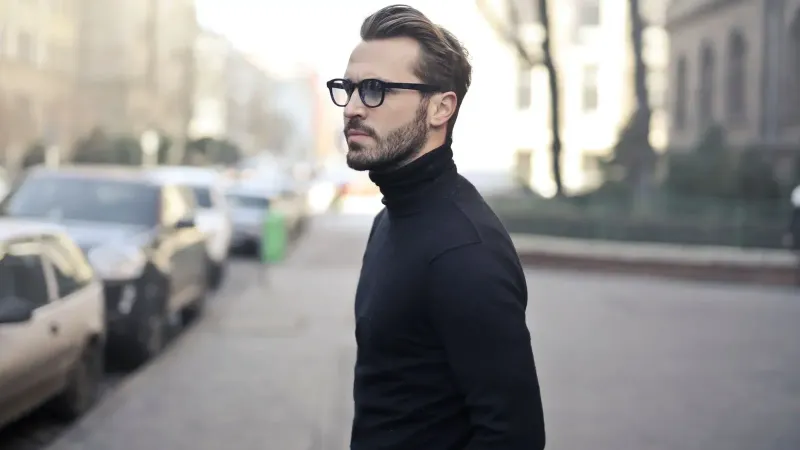 A fashionable man in a black top and glasses poses in an urban street with parked cars.
