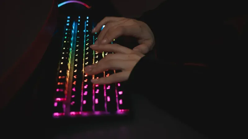 Close-up of hands typing on a colorful RGB backlit mechanical keyboard in a dimly lit environment.