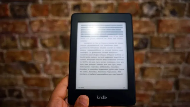 Close-up of a Kindle e-reader held by a person, showcasing a digital reading device against a textured brick wall backdrop.