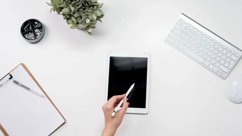 A minimalist home office setup featuring a tablet, stylus, keyboard, plant, and stationery on a white desk.