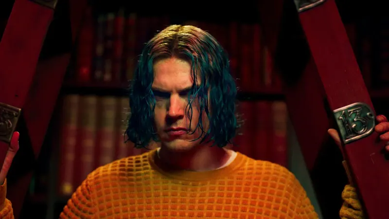 Kai Anderson (Evan Peters) mira hacia adelante con una expresión intensa, enmarcado por una estructura roja con una placa de metal que muestra el número 13; usa un suéter naranja de punto
