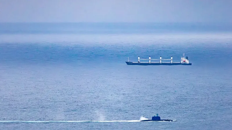 Un barco de carga y un submarino navegan por el vasto océano abierto bajo un cielo despejado.