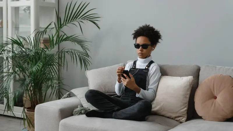 Un adolescente con discapacidad visual sentado en un sofá usando un smartphone y auriculares.