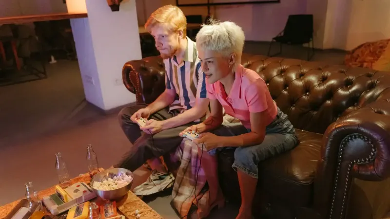 Two friends playing video games on a leather sofa, surrounded by snacks and drinks, enjoying a casual gaming night.