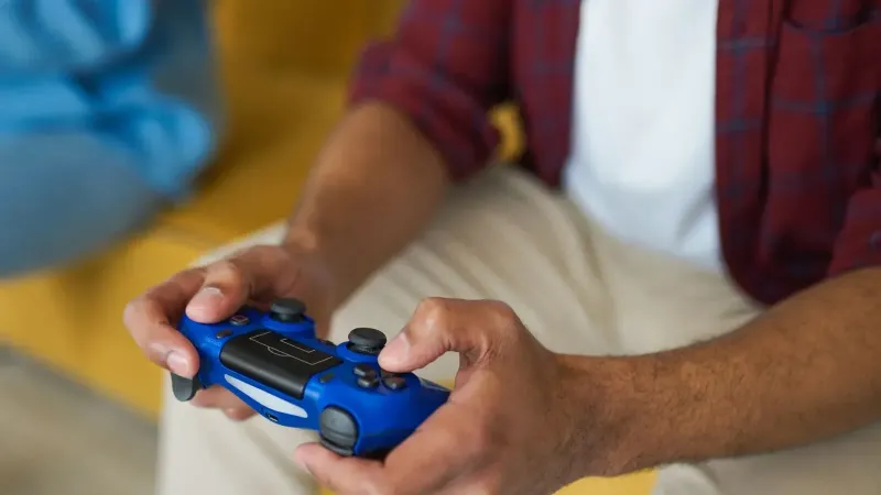 Manos sosteniendo un controlador de juego inalámbrico azul en un cómodo sofá en el interior.