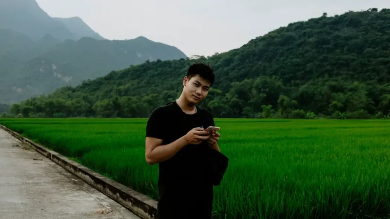 Hombre asiático de pie con smartphone en un paisaje rural brumoso, rodeado de campos de arroz verde y montañas.