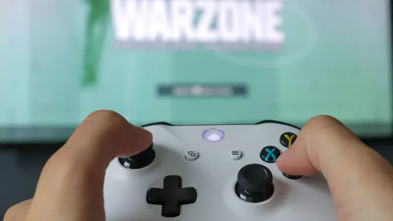 Un primer plano de manos sosteniendo un controlador Xbox contra una pantalla de juego, destacando la experiencia de juego.
