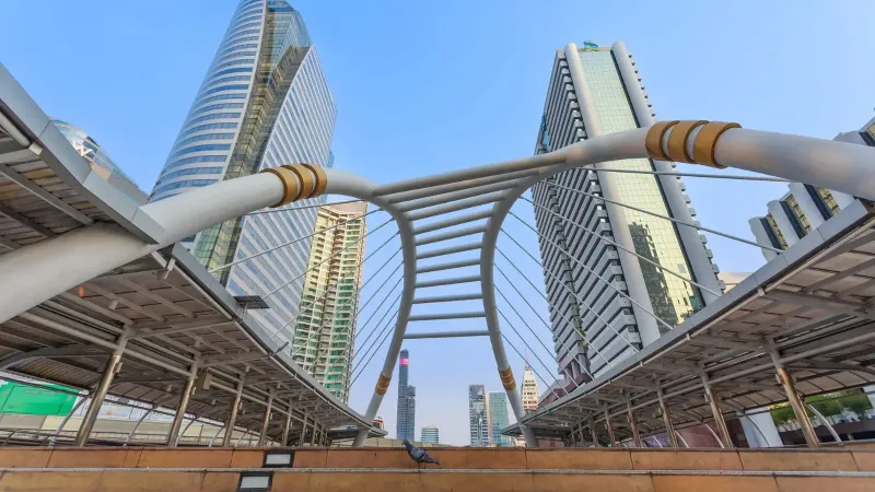Vista dinámica del horizonte de Bangkok con imponentes rascacielos y el icónico puente Skywalk bajo un cielo azul claro.