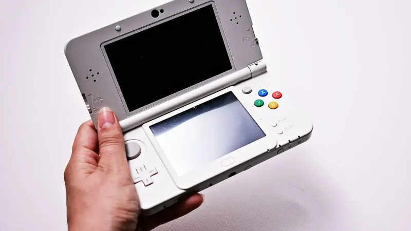 Primer plano de una mano que sostiene una consola de juegos portátil abierta, con pantallas duales.