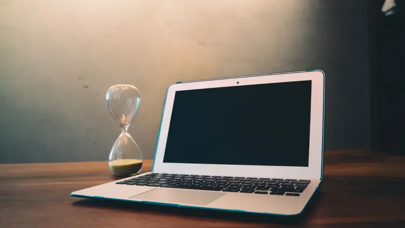 Modern laptop on a wooden desk beside an hourglass, symbolizing time and technology intersection.