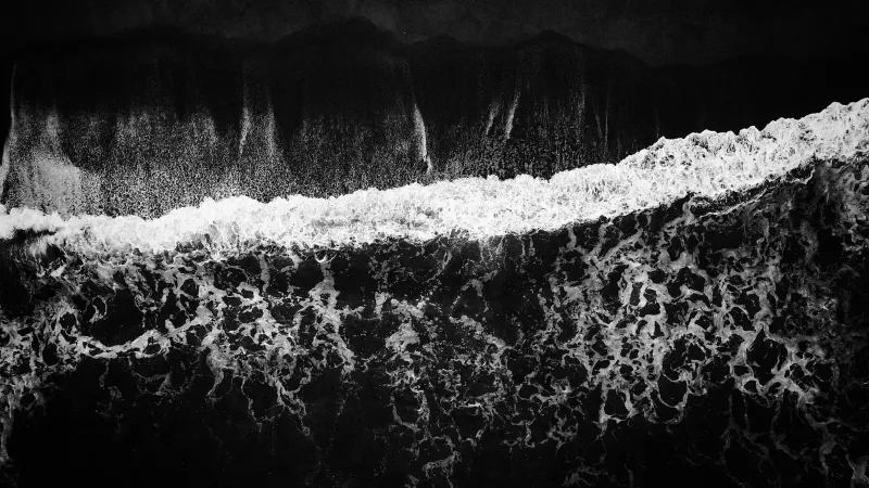 Vista de pájaro de las olas del océano chocando en una orilla oscura, capturada en blanco y negro.