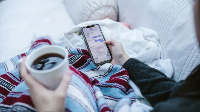 Persona que se relaja en el interior con café y smartphone, enviando mensajes de texto en un ambiente acogedor.