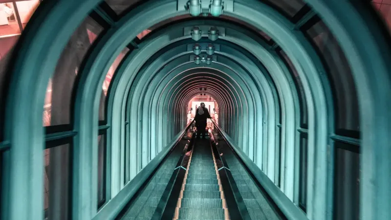 Un túnel futurista con una escalera mecánica iluminada y siluetas de personas, mostrando la arquitectura moderna.