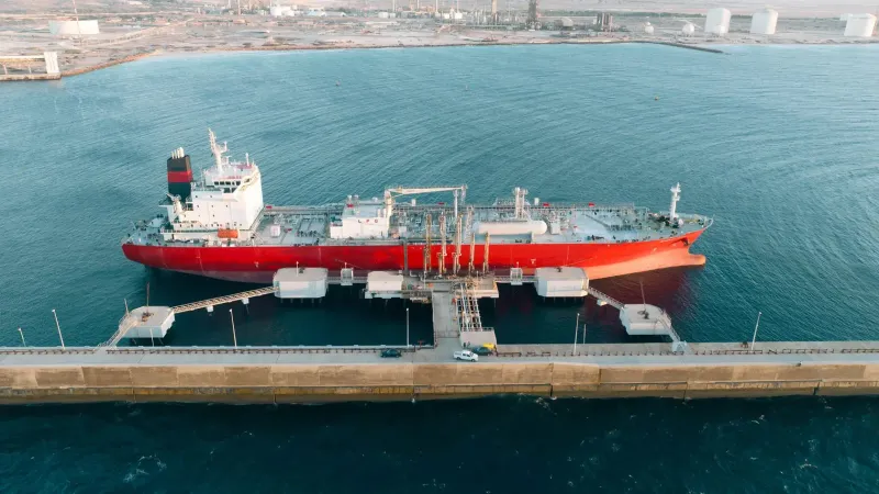 Disparo aéreo de un buque de carga rojo atracado en un puerto industrial con agua azul claro.