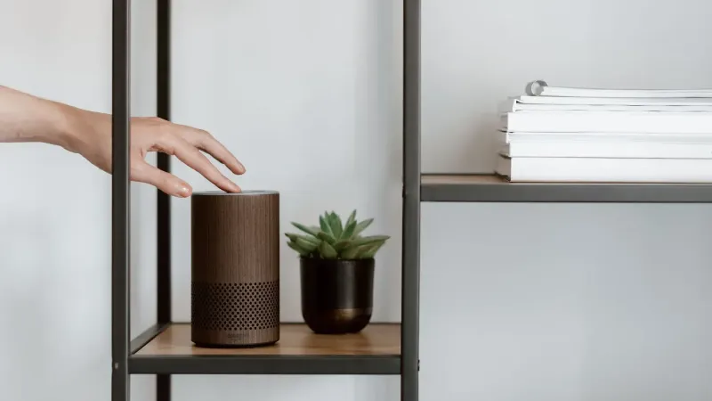 Una mano interactúa con un altavoz inteligente en un estante minimalista, con una planta y libros apilados.