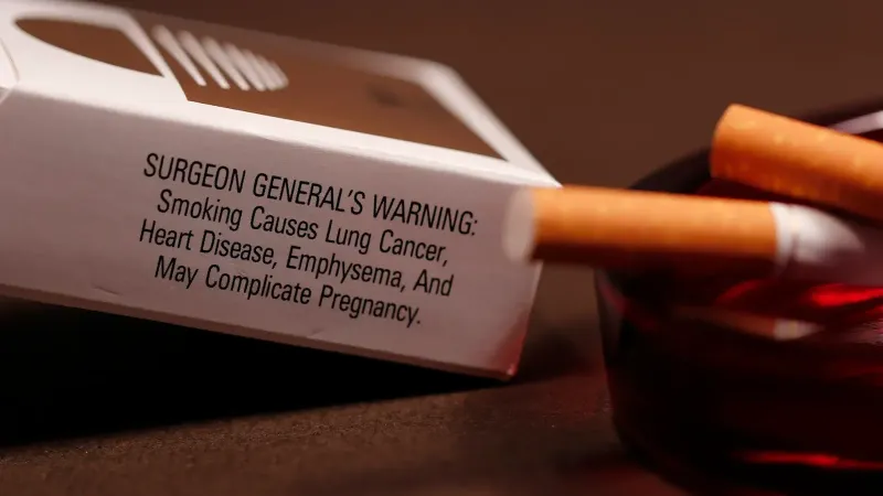 En su trasfondo, esta imagen muestra un paquete abierto de cigarrillos con su advertencia de salud en el panel lateral a los posibles fumadores que indican algunos de los efectos nocivos