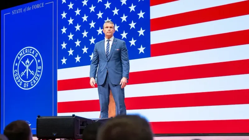 A person in business attire stands on a stage, with a large American flag in the background; people are seated in front of the stage.